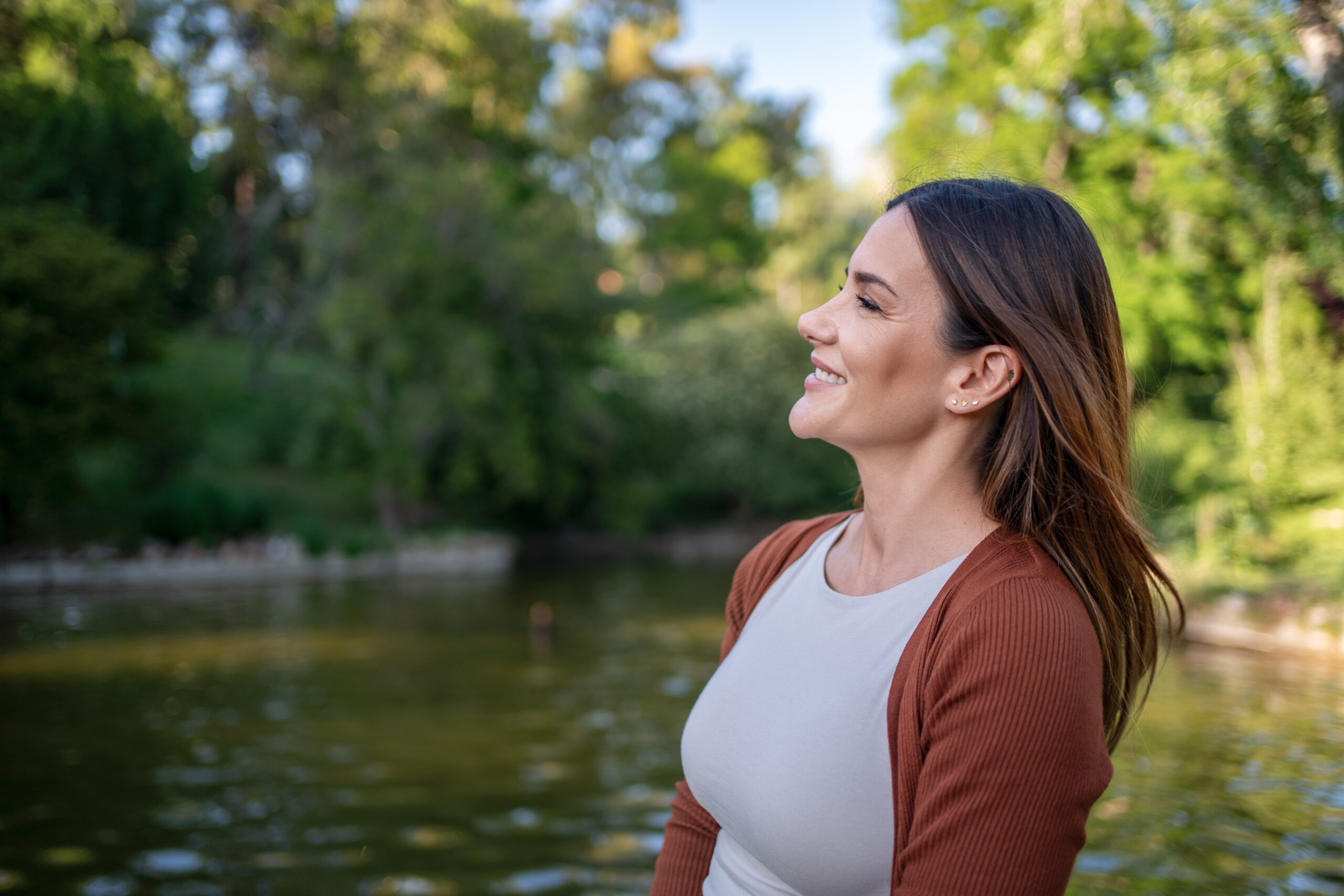 Smiling woman in white top and brown cardigan by a pond in a park.
