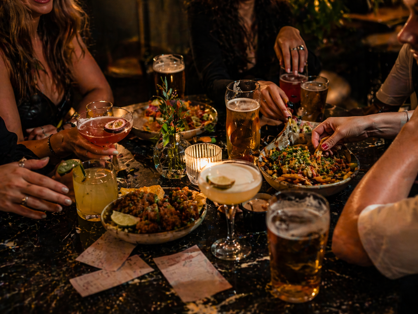 A bar table filled with cocktails, pints, loaded sharing plates and karaoke song slips