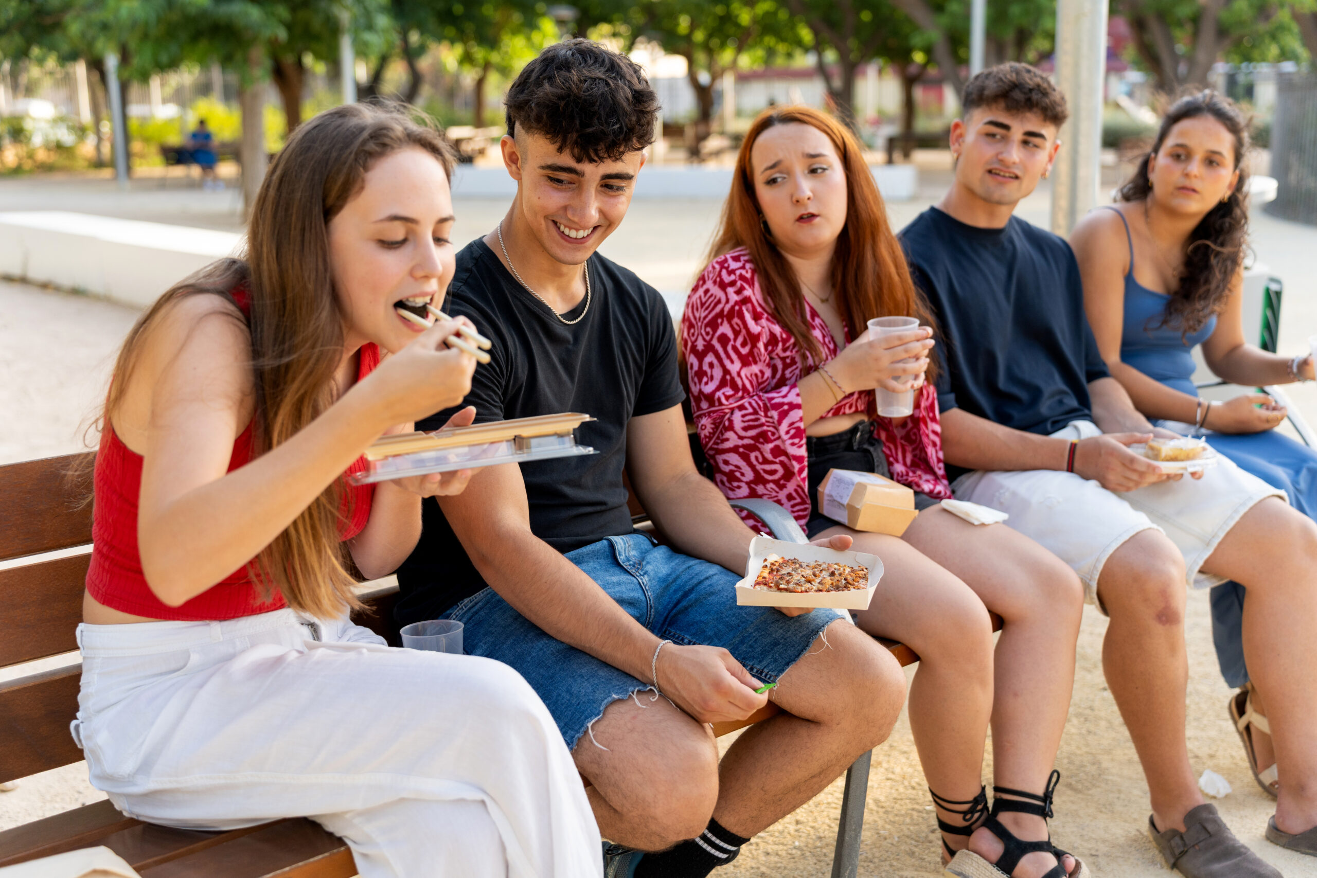 Five teenager friends sitting on a bench, eating take away food and drinking, enjoying their time together