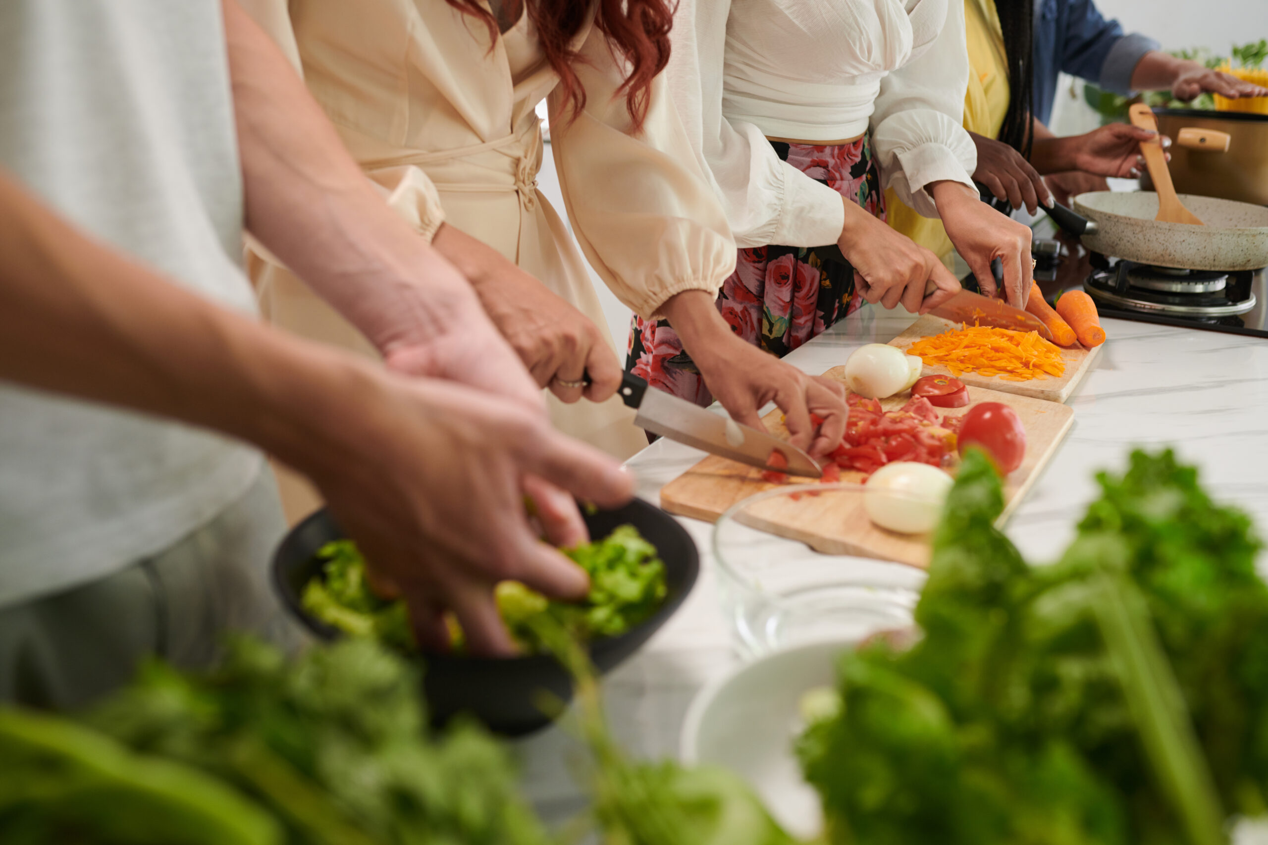 Row of young friends with sharp knives chopping fresh vegetables for salad while standing by table and cooking dinner