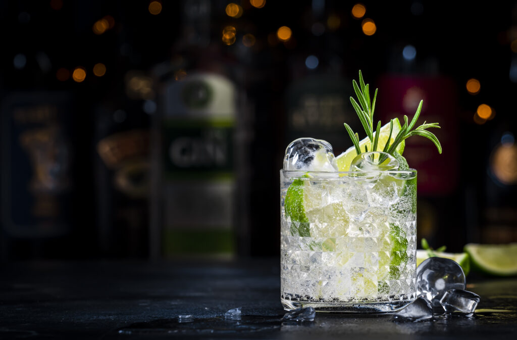 Gin fizz cocktail drink with dry gin, lime juice, sugar syrup, soda, fresh rosemary and ice. Black bar counter background, steel bar tools and bottles