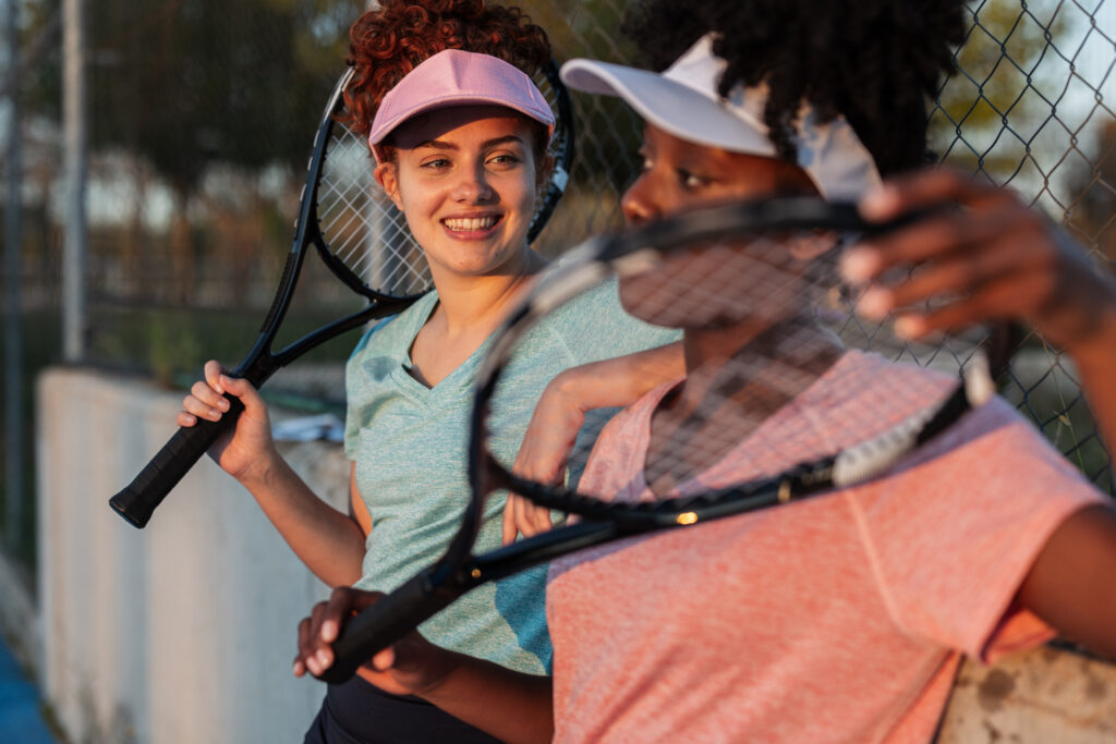 Two joyful multi ethnic female tennis players enjoying a moment of relaxation after an exciting match, holding their rackets and soaking in the warm sun on a beautiful afternoon